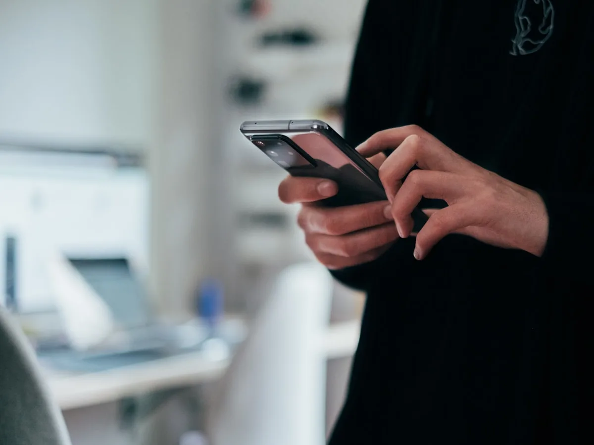 A pair of hands holding a phone at a desk, with a laptop out of focus behind them.